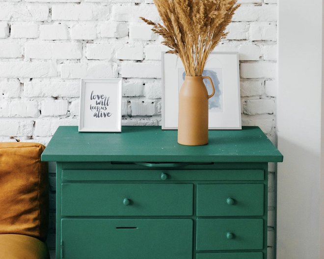 A green dresser with a vase and a picture frame on top, against a white brick wall